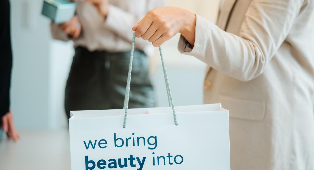 BEAUTY SOLUTIONS Branding & Merchandise | Tragtasche VIVABEAU | © BEAUTY SOLUTIONS | Martin Hofmann Fotografie Eine Frau hält eine hochwertige Papier-Einkaufstasche mit der Aufschrift 'We bring beauty into your life' von VIVABEAU. Im Hintergrund sind weitere Personen zu sehen, die sich in einer modernen Umgebung unterhalten. Die Szene vermittelt ein stilvolles Shopping-Erlebnis.